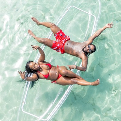 Two people in swimwear relax on a clear kayak in turquoise water, seen from above.