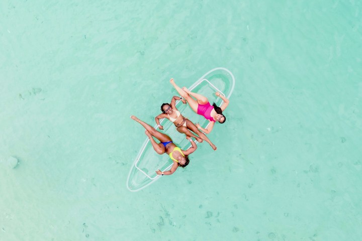 Three people in swimwear lounging on a transparent float in clear turquoise water.