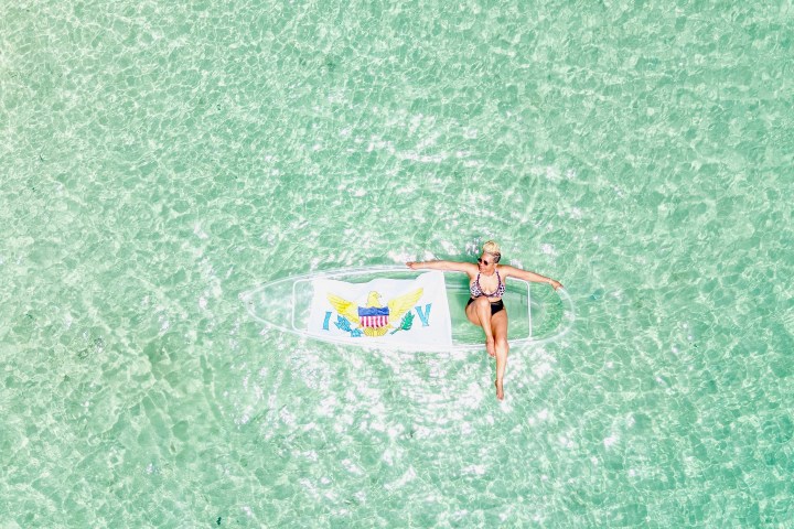 Aerial view of person in swimwear floating on a transparent kayak with a flag in clear turquoise water.