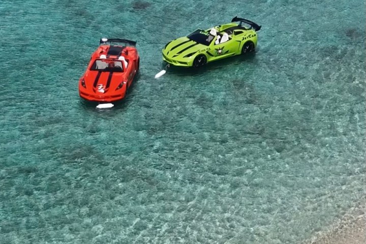 Two car-shaped jet skis, one red and one green, float on clear turquoise water near a sandy shore.